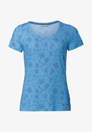 Mujer con una camiseta azul de flores de manga corta, pantalones cortos negros y zapatillas azules, de pie con una mano en el bolsillo.