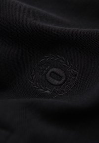 Schwarz strukturiertes Gewebe mit subtil besticktem kreisförmigem Emblem mit dem Buchstaben "O" und umgebenden dekorativen Elementen.