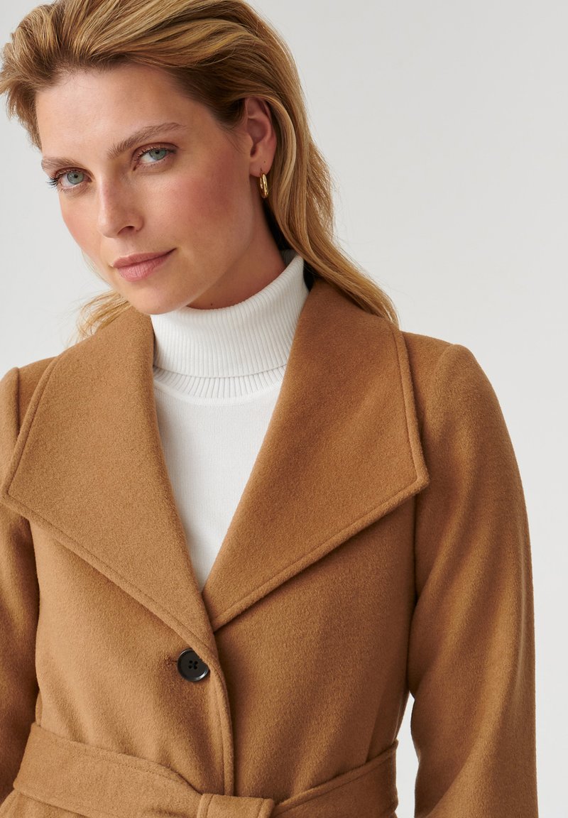 Cappotto di lana marrone con ampio colletto, chiusura a un unico bottone nero, vita con cintura e texture morbida, abbinato a un dolcevita bianco sotto.