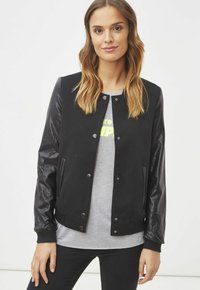Chaqueta bomber negra con mangas de cuero, botones de presión, puños acanalados y bolsillos laterales. Usada sobre una camiseta gris con un diseño impreso.