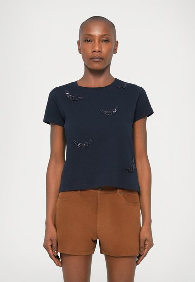 Marineblaues Baumwoll-T-Shirt mit kurzen Ärmeln und Rundhalsausschnitt, verziert mit schwarzen gestickten Halbmondformen auf der Vorderseite. Kombiniert mit braunen Shorts.