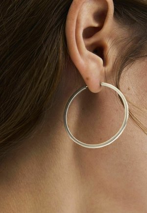 Primer plano de la oreja de una persona con cabello castaño claro que lleva un gran pendiente de aro fino plateado.