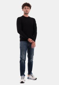Maglione di lana nera, jeans slim-fit di colore blu scuro e sneakers sportive bianche con dettagli blu e arancioni. Il modello è in piedi con le mani rilassate.