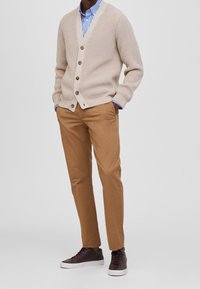 Beżowy dziergany kardigan z sześcioma guzikami na niebieskiej i białej w paski koszuli. Brązowe chinosy i ciemne sneakersy dopełniają stylizację.