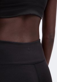 Gros plan d'une personne portant un legging noir taille haute et un crop top assorti, montrant le ventre et un petit logo sur le legging.