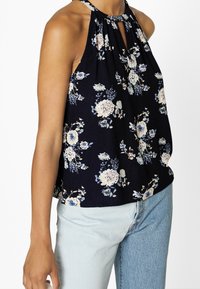 Haut halter floral en tissu bleu marine foncé, arborant des fleurs blanches et bleu clair, avec un décolleté en forme de clef et une coupe ample, associé à un jean bleu clair.