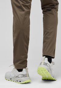 Lichtgrijze sportschoenen met textuurpatronen, een dikke zool in limoengroen en reflecterende accenten, gecombineerd met bruine tapered broeken.