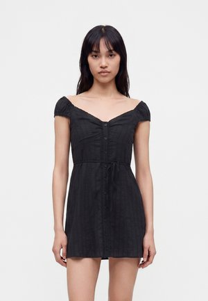 Femme portant une courte robe noire épaules dénudées avec des boutons sur le devant et un détail noué à la taille, debout devant un fond uni.