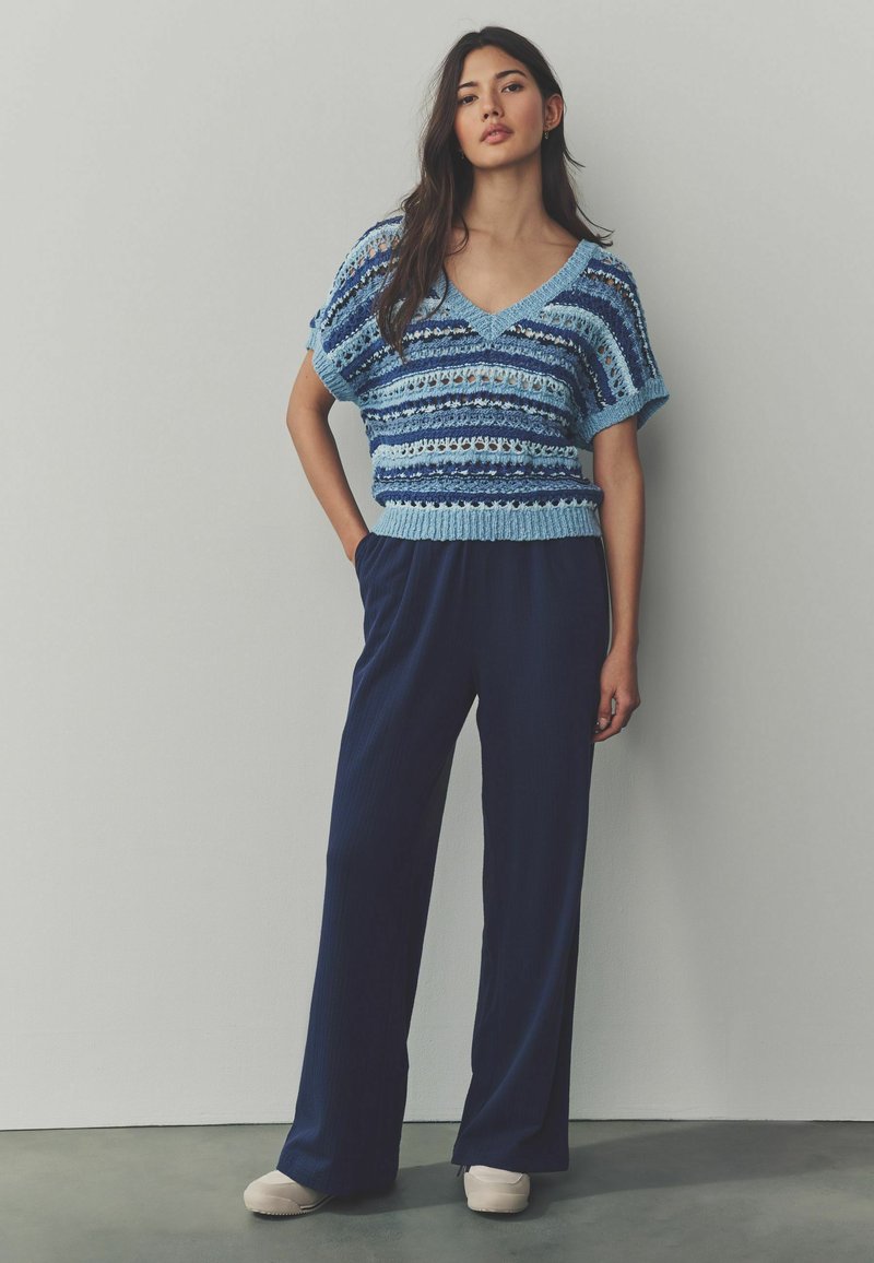 Pulover deschis la culoare, din tricot cu model deschis, cu dungi orizontale și un decolteu în V larg, asortat cu pantaloni plisati bleumarin și pantofi de culoare crem.