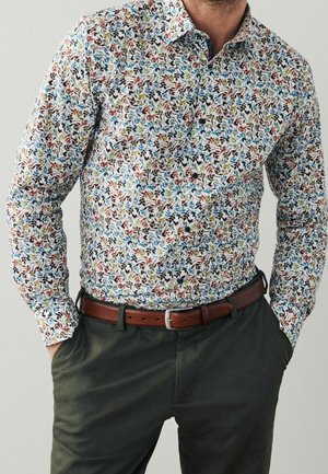 Homme portant une chemise à manches longues à motifs floraux, avec une ceinture en cuir marron et un pantalon vert foncé, les mains dans les poches.