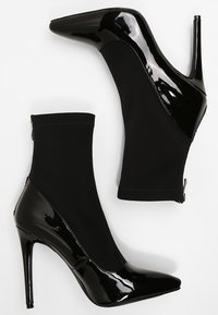 Bottines noires à talons hauts avec bouts pointus, zones du bout et du talon en cuir verni brillant, et tige en tissu extensible.