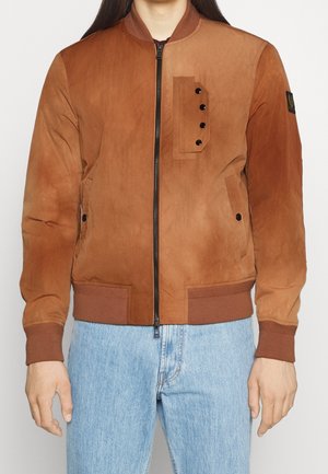 Mężczyzna w brązowej bomberce ze skóry zamszowej z czarnymi zatrzaskami na kieszeni na piersi i jasnoniebieskich dżinsach, stojący na tle jednolitego tła.