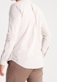 Camisa beige claro de manga larga con dobladillo redondeado, puños con botones y detalle de pliegue en la parte trasera. Confeccionada con un tejido texturizado.