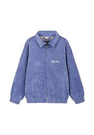 Young woman with long hair leans on a bus seat inside an empty bus with black leather seats and overhead luggage racks.