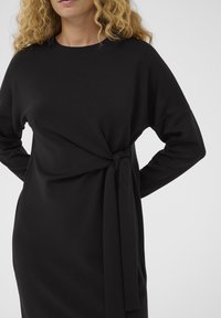 Kobieta z kręconymi blond włosami, ubrana w czarną sukienkę z długim rękawem związaną na wstążkę w talii, na jednolitym tle.