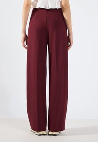 Burgundy kalhoty s volným střihem z hladkého materiálu, s ostrými záhyby a vysokým pasem, nošené s lehkou obuví.