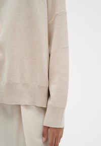 Pull en tricot beige à coupe décontractée, épaules tombantes et poignets côtelés. Texture douce et design décontracté, associé à un tissu de couleur claire en dessous.