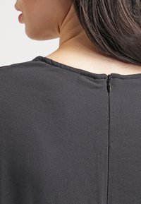 Musta, teksturoitu kangastoppi, jossa on takatasku, pyöreä kaula-aukko ja suora siluetti, joka laskeutuu hartioille.