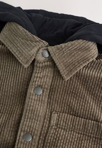 Veste en velours côtelé marron avec une capuche noire, présentant une texture côtelée, des boutons-pression et une poche avant.