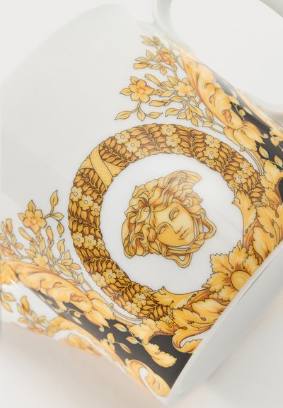 Λευκή κεραμική κούπα με χρυσό λουλουδάτο και σπειροειδές σχέδιο που περιβάλλει ένα κεντρικό έμβλημα με κεφάλι Μέδουσας.