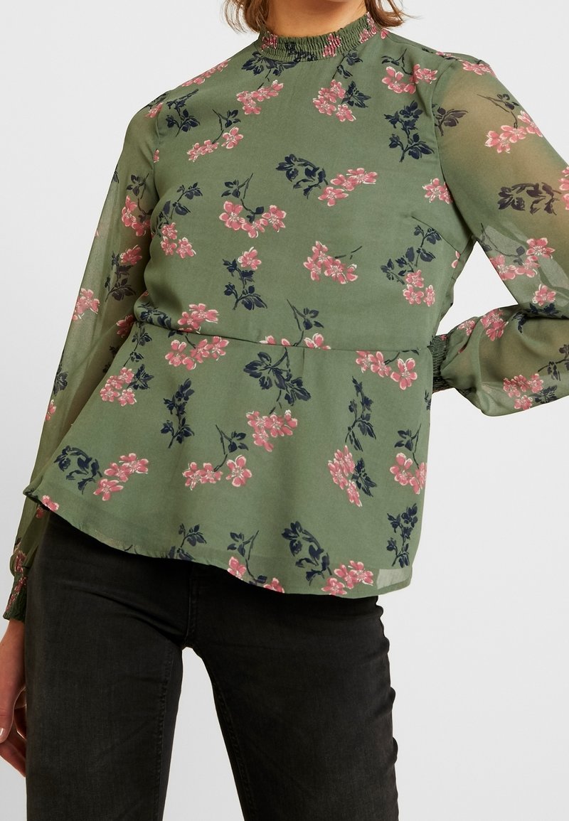 Green floral blouse with long sleeves, featuring a smocked neckline, pink flowers, and a peplum silhouette in lightweight fabric.