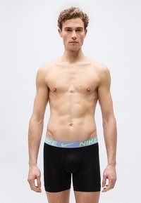 Νεαρός άνδρας που στέκεται στραμμένος προς τα εμπρός, φοράει μαύρα εσώρουχα boxer της Nike με φαρδιά μέση σε νεον πράσινο και γκρι, ουδέτερη έκφραση, απλό ανοιχτό φόντο.