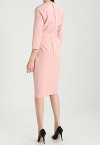 Robe ajustée rose avec une fermeture éclair dans le dos, des manches trois-quarts et une taille ceinturée. Le mannequin porte des escarpins noirs.