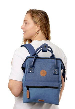 Junge Frau mit welligem Haar, die ein weißes Hemd und einen blauen rechteckigen Rucksack mit schwarzem Reißverschluss und Lederakzenten trägt.