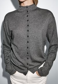 Cardigan gris en maille avec un col montant, présentant une fermeture à boutons et des manches longues. Le tissu a une texture douce et une coupe décontractée.
