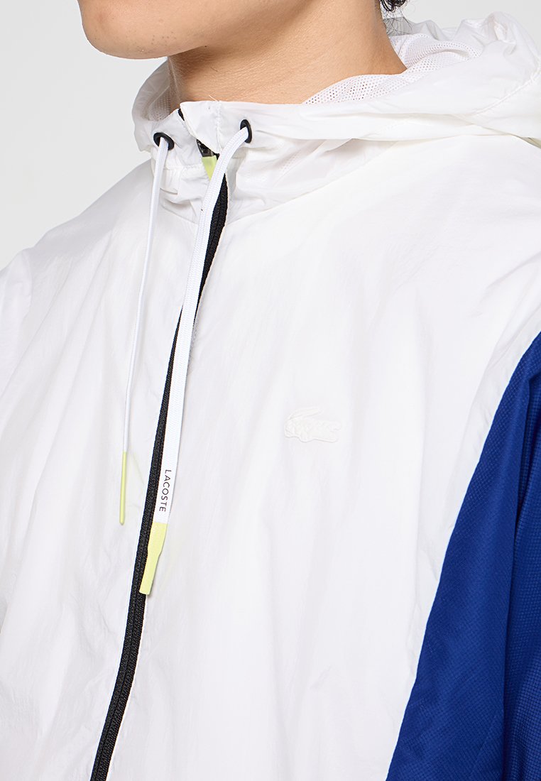 Veste coupe-vent blanche et marine avec capuche à cordon, tissu texturé et petit logo en relief sur la poitrine. Présente des détails de fermeture éclair noirs.