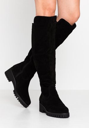 Bottes montantes en daim noir avec semelles épaisses à reliefs et talons carrés portées jambes croisées sur un fond clair uni.
