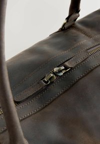 Bruine leren tas met een gladde textuur, voorzien van een opvallende rits met metalen hardware, en contrasterende stiksels langs de naden.