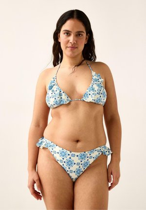 Pitkähiuksinen nainen, jolla on valkoinen sinikukkakuviollinen bikini ja röyhelöyksityiskohdat, seisoo yksinkertaisen vaalean taustan edessä.