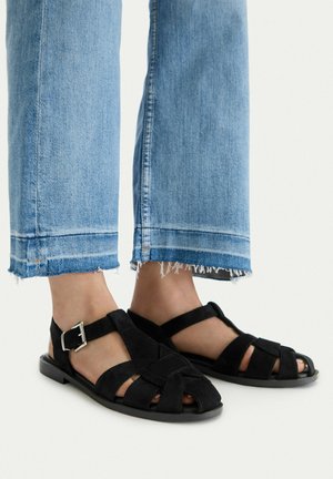 Nogi w krótkich niebieskich dżinsach, noszące czarne sandały z zakrytymi palcami i paskami z klamrami na białym tle.