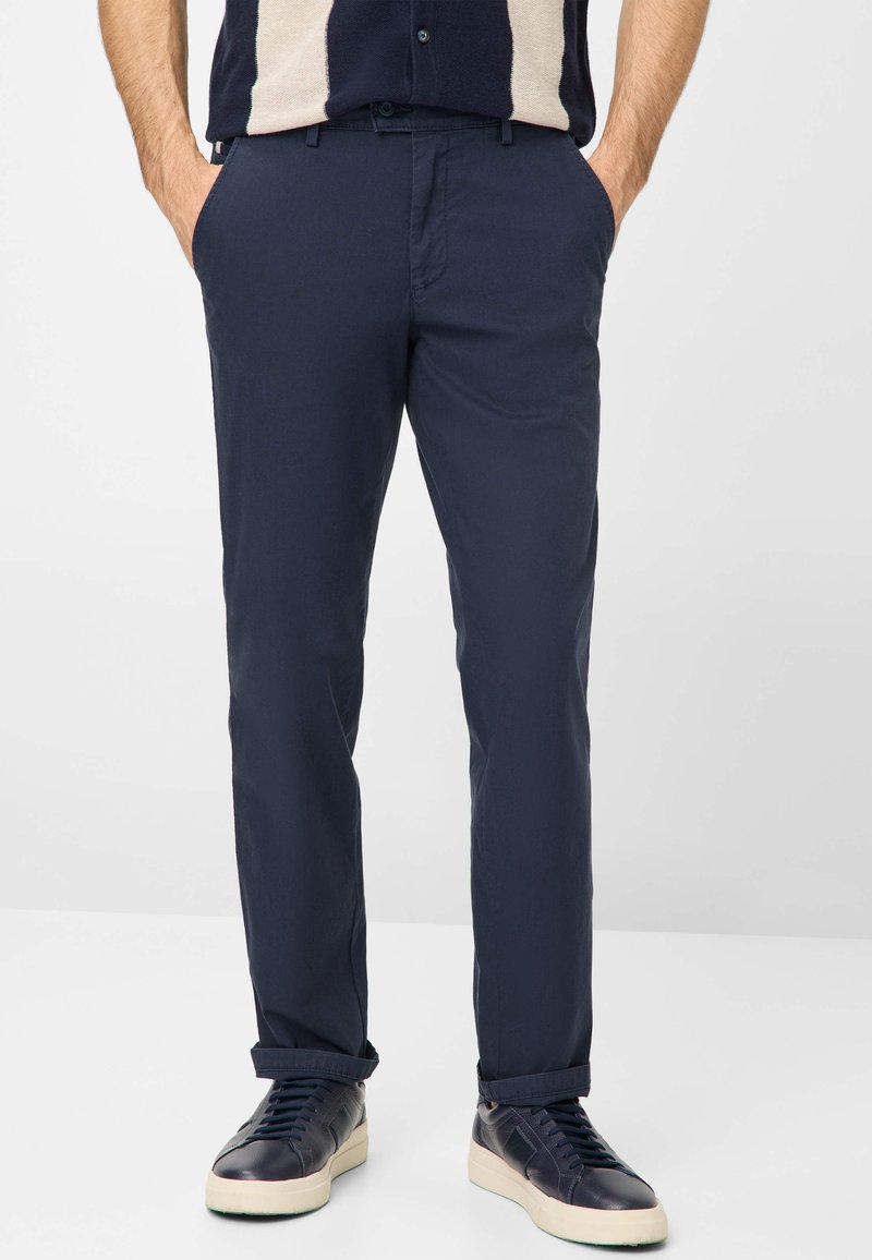 Navy blue chinos with a smooth texture, featuring side pockets and rolled cuffs, paired with dark sneakers. Simple design and modern fit.