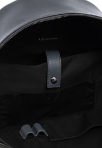 Zaino con parte superiore arrotondata, realizzato in materiale grigio scuro testurizzato. L'interno presenta una fodera nera, una tracolla in pelle con chiusura a scatto e tasche.