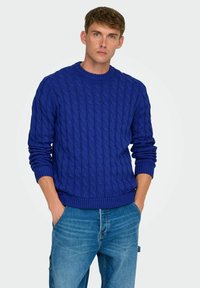 Blå kabelstrikket sweater med rund halsudskæring og ribstrikkede manchetter, parret med lyseblå denimjeans. Glat tekstur og afslappet pasform.