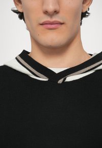 Fekete kötött pulóver V-nyakkivágással és fehér kapucnival, amelyen szürke és fekete csíkok találhatók. Az anyag texturált és testhez simuló hatású.