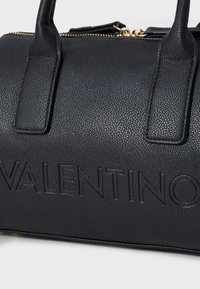 Sac à main en cuir noir avec logo embossé, forme structurée, deux poignées supérieures et détails de fermeture éclair dorés. Surface avec un motif texturé.