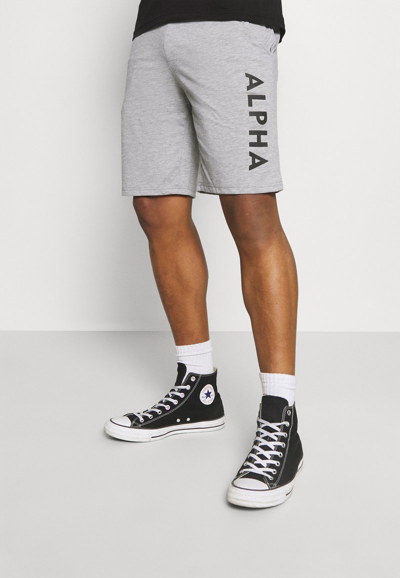 Shorts de sport gris en mélange de coton, arborant une impression latérale "ALPHA" en noir. Associés à des baskets montantes noires et des chaussettes blanches.
