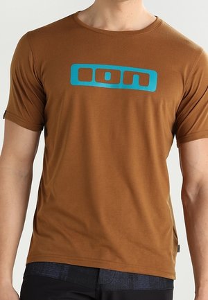 Mężczyzna noszący brązowy T-shirt z krótkim rękawem z niebieskim logo "ion" na środku klatki piersiowej.
