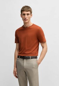 T-shirt a maniche corte di colore ruggine con una finitura texture. Abbinato a pantaloni beige chiaro su misura e una cintura marrone scuro.
