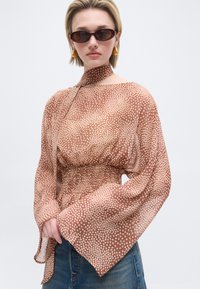 Γυναίκα που φοράει μπλούζα με καφέ και μπεζ πουά και μεγάλα μανίκια, μπλε τζιν, χρυσά σκουλαρίκια και σκούρα οβάλ γυαλιά ηλίου σε απλό φόντο.