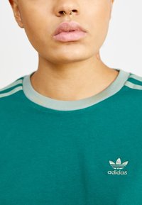 Teal színű pamut póló, bordázott kerek nyakkal, három függőleges csíkkal a vállakon és hímzett Adidas logóval.
