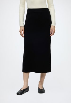 Femme portant une jupe midi noire associée à un haut côtelé crème à manches longues et des chaussures plates noires sur fond blanc.