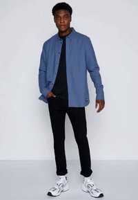 Light blue button-up shirt over a black t-shirt, paired with black pants and white sneakers. Model stands against a plain background.