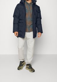 Marineblå puffjakke med hætte, gråternet skjorte under, lysegrå sweatpants og grønne løbesko med blå accenter.