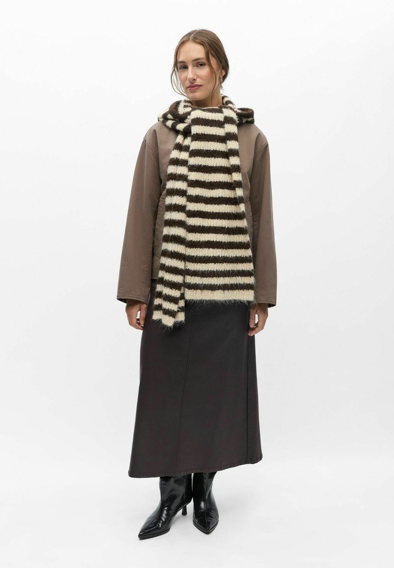 Femme portant une veste marron, une longue jupe noire, des bottes noires pointues et une épaisse écharpe rayée noir et blanc sur un fond blanc.