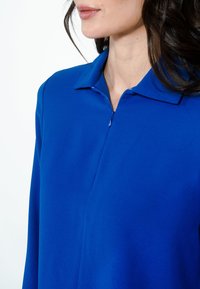Egy nő royal kék, hosszú ujjú, galléros felsőt visel, amely elöl zipzárral záródik, a vállától az álláig mutatva, fehér háttér előtt.