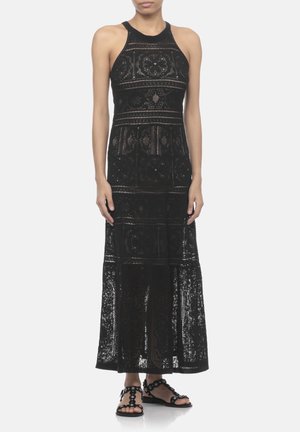 Donna che indossa un abito maxi nero senza maniche all'uncinetto con motivi floreali abbinato a sandali piatti neri decorati con borchie circolari.